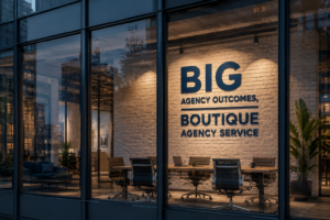 Conference room inside a glass-walled office with a brick wall featuring the sign 'BIG AGENCY OUTCOMES, BOUTIQUE AGENCY SERVICE'.